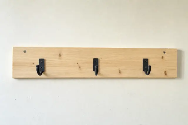 Single pine board coat rack with three black hooks mounted on an apartment hallway wall, snapshot from the doorway